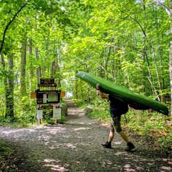 A man carries a kayak past signs pointing to 13th lake in the summer with green light filtering through the Summer canopy.